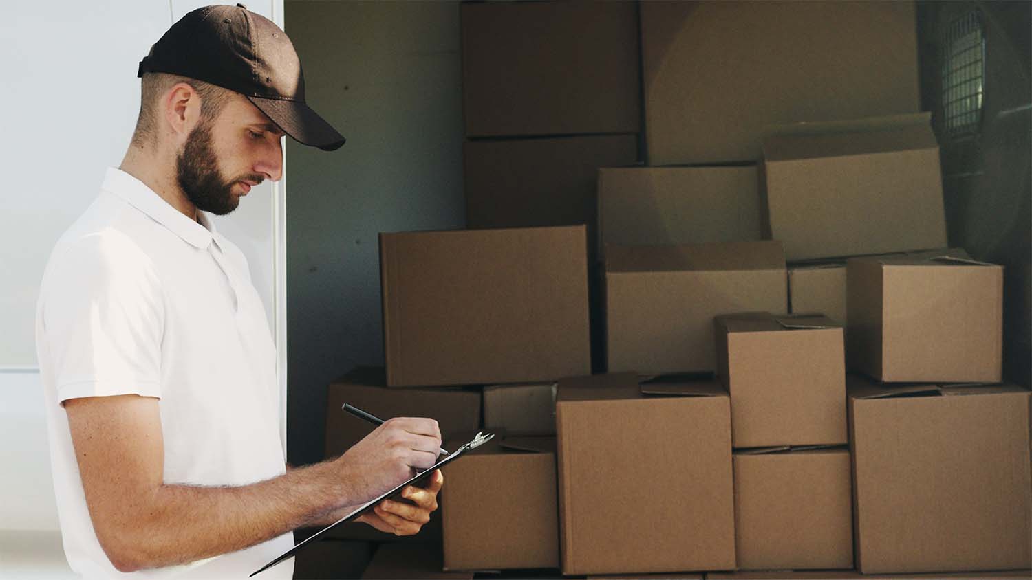 A delivery professional writing on a clipboard standing in front of a truck full of packages to be delivered